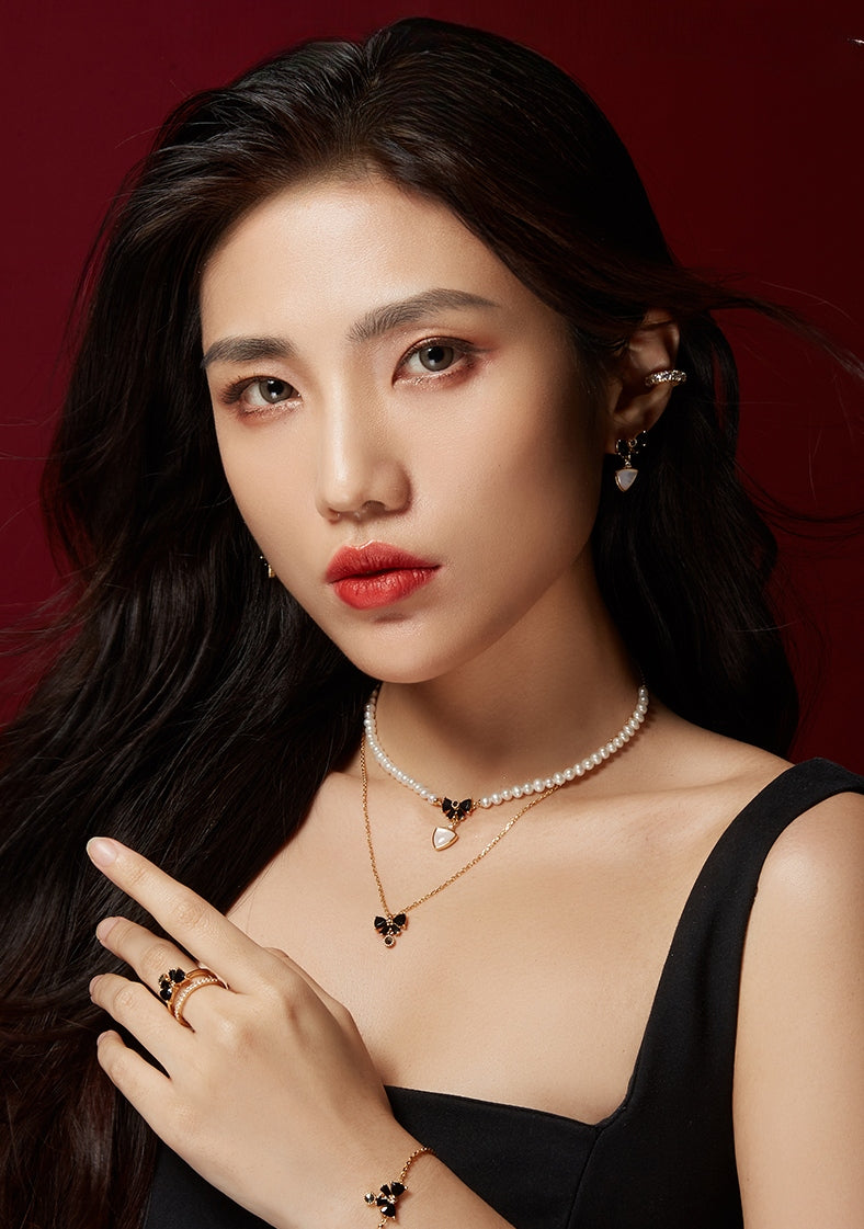 A woman showcases elegant jewelry, featuring pearl and gold butterfly necklaces, rings, and earrings against a deep red background.