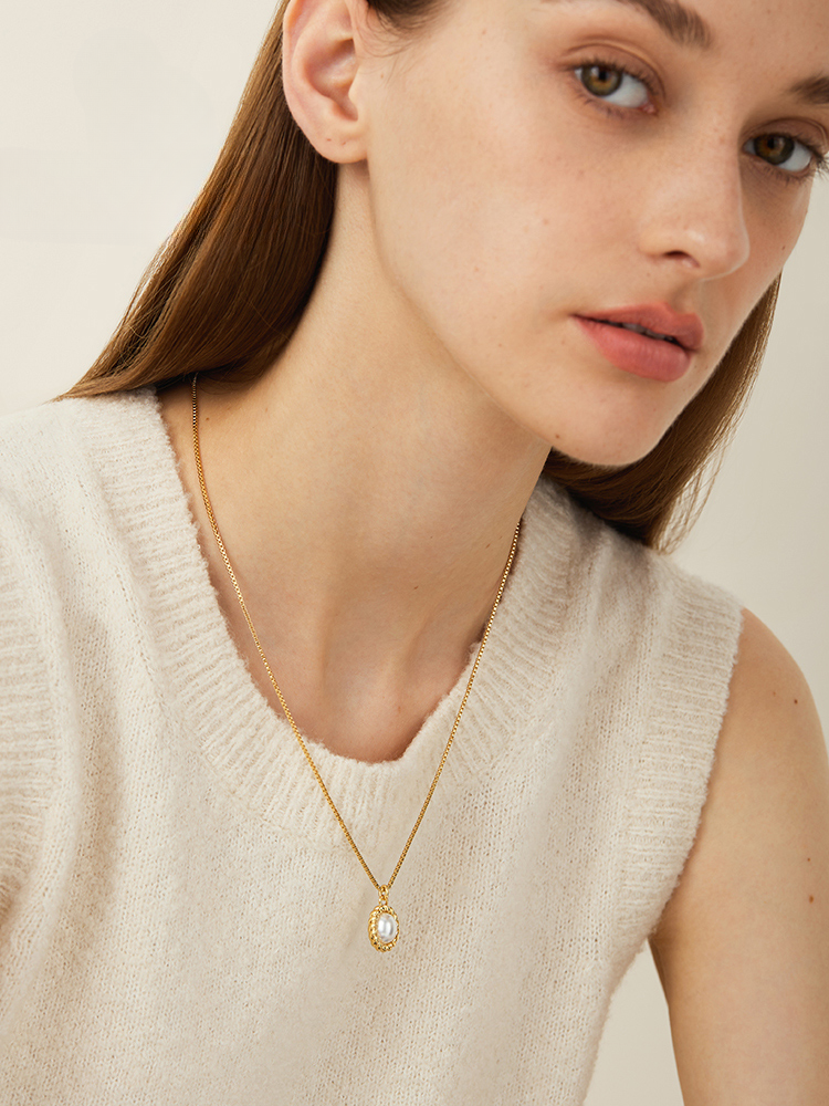 A close-up of a person wearing a cream sleeveless sweater and a delicate gold necklace featuring an oval pendant with a shimmering stone.
