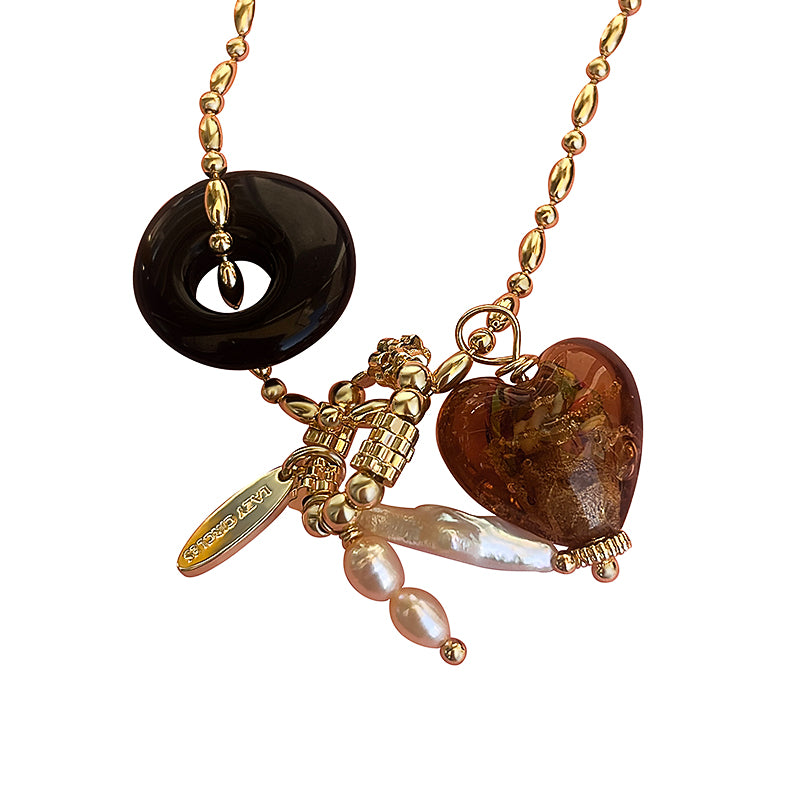 Close-up of a gold necklace adorned with a black donut pendant, a brown heart charm, and lustrous pearl accents.