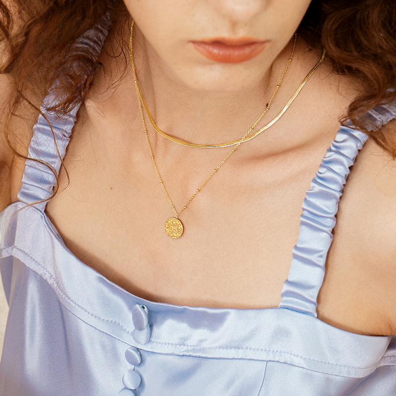 A close-up of a person's neck and chest wearing layered gold necklaces, with a focus on a round pendant, against a light blue satin top.