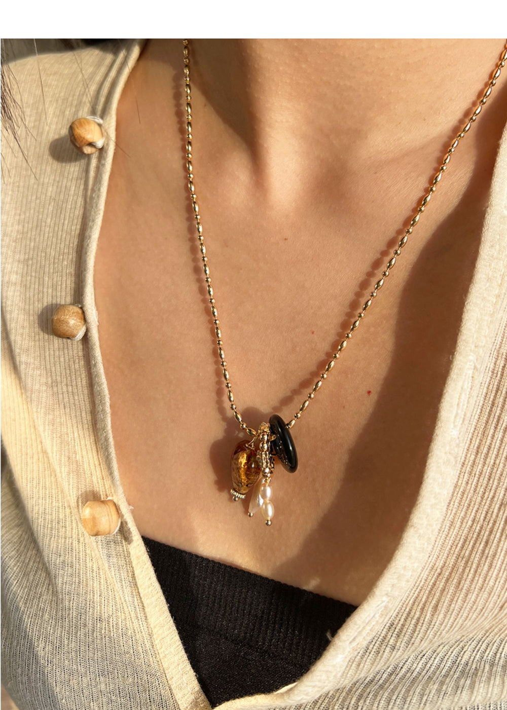 A golden chain necklace featuring a mix of charms, including a shell, a black button, and a pearl, worn against soft fabric.